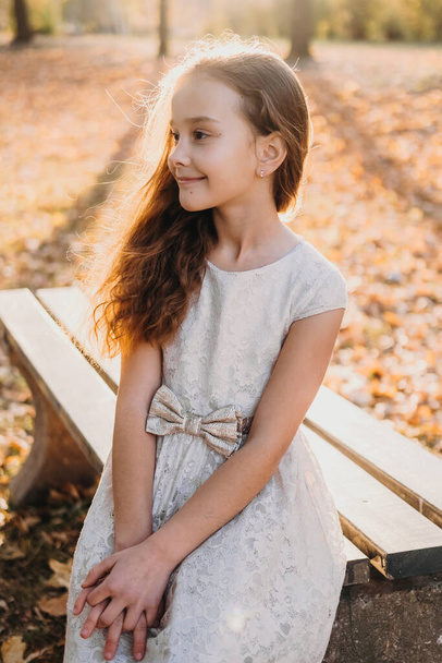 Портрет симпатичной маленькой девочки, расслабляющей в красоте осенний пейзаж в белом платье, сидящей на скамейке в осеннем парке. Наружный образ жизни. Улыбающаяся женщина на улице - Фото, изображение