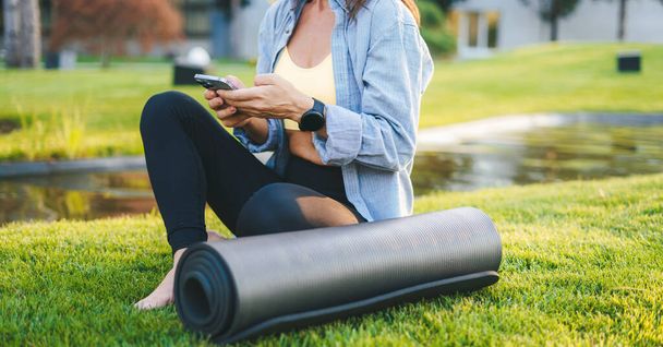 Gehakt beeld van een vrouw kijken naar video op smartphone zitten op gras oppervlak, genieten van 's morgens tijd. Vrouw die haar smartphone gebruikt om een sms te sturen. Sportief van dichtbij - Foto, afbeelding