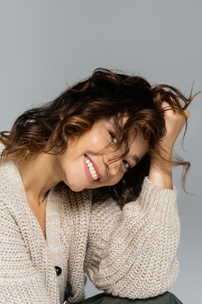 glückliche Frau in Strickjacke berührt welliges Haar und blickt vereinzelt in die Kamera auf grau - Foto, Bild