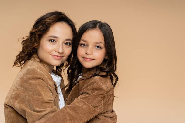 mom and daughter in stylish suede jackets smiling at camera while hugging isolated on beige - Fotoğraf, Görsel