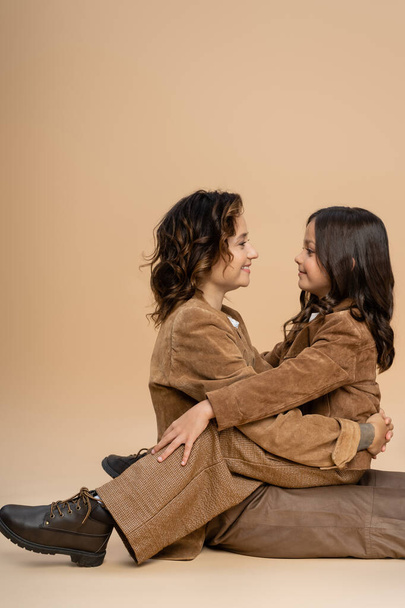side view of mom and daughter in stylish autumn outfit hugging and smiling at each other on beige background - Foto, Imagem