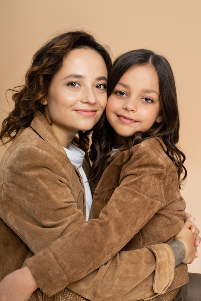brunette mom and daughter in brown autumn jackets hugging and smiling isolated on beige - Photo, Image