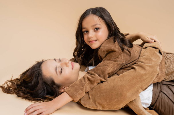 stylish girl smiling at camera near mom lying with closed eyes on beige background - Foto, imagen