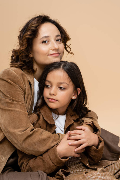 mujer feliz y elegante abrazando hija en chaqueta de gamuza marrón aislado en beige - Foto, imagen