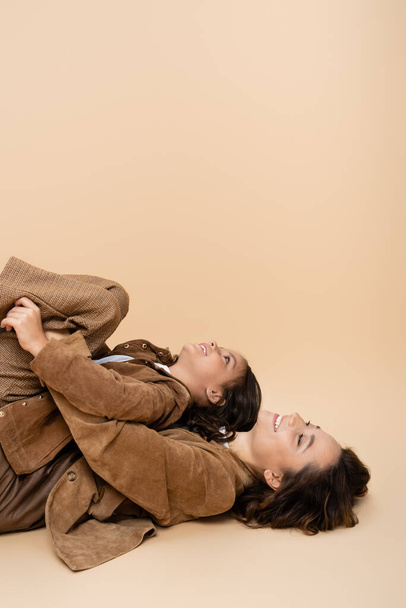 side view of girl having fun and lying on happy mom in autumn clothes on beige background - Photo, image