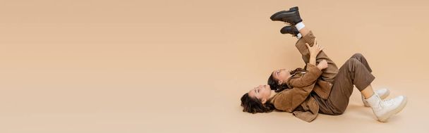 side view of girl in trendy boots lying on mom with raised legs while having fun on beige background, banner - Fotó, kép