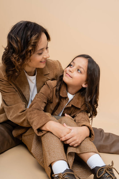 joyful woman and child in stylish suede jackets looking at each other on beige background - Фото, изображение