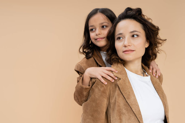 brunette girl hugging shoulders of mom in brown suede jacket while looking away isolated on beige - Фото, изображение