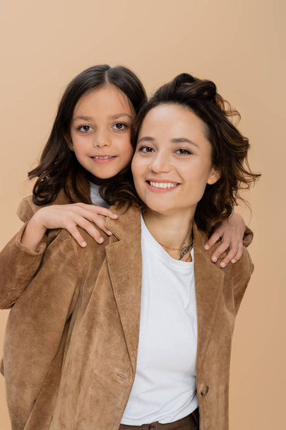 brunette girl hugging shoulders of happy mom in brown suede jacket isolated on beige - Photo, Image