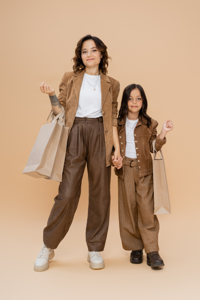 full length of woman and girl in stylish autumn clothes standing with shopping bags on beige background - Fotografie, Obrázek