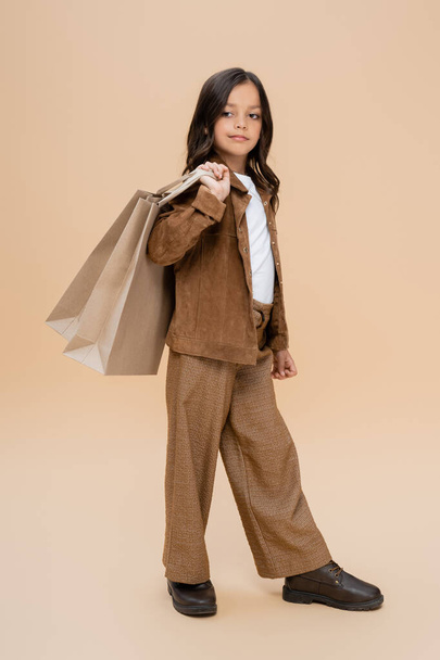 full length of girl in suede jacket and brown pants holding shopping bags on beige background - Φωτογραφία, εικόνα