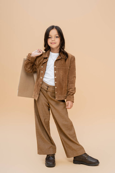 brunette meisje in suède jas en bruine broek poseren met boodschappentassen op beige achtergrond - Foto, afbeelding