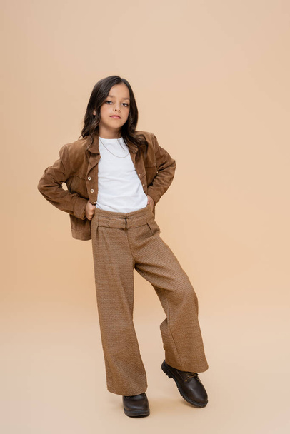 full length of trendy girl in brown pants and suede jacket standing with hands on hips on beige background - Фото, изображение
