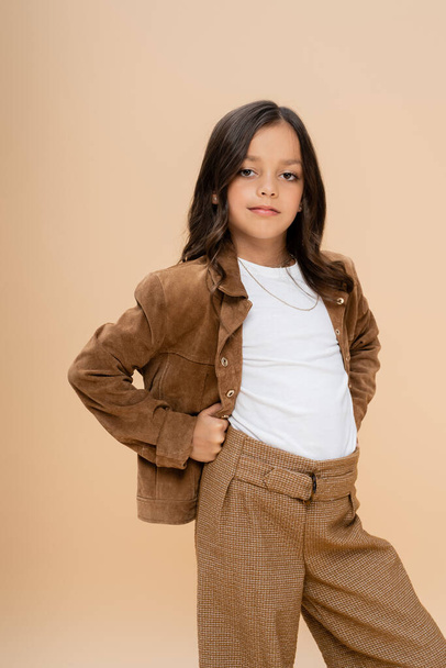 brunette girl in brown suede jacket posing with hands on hips while looking at camera isolated on beige - Fotografie, Obrázek