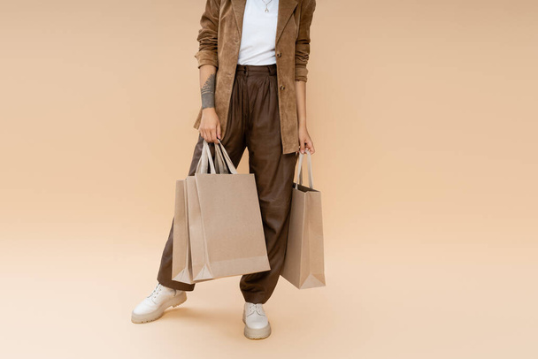 partial view of tattooed woman in brown pants holding shopping bags on beige background - Φωτογραφία, εικόνα