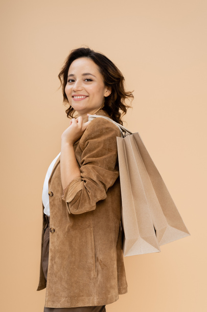 brunette woman in brown suede jacket holding shopping bags while smiling at camera isolated on beige - Foto, imagen