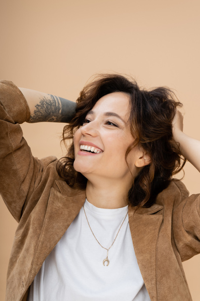 happy tattooed woman in brown suede jacket touching wavy hair and looking away isolated on beige - Photo, Image