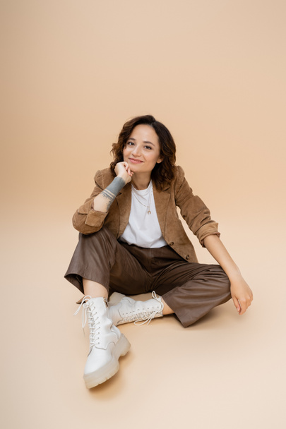 brunette woman with wavy hair wearing trendy autumn clothes while sitting with hand near face on beige background - Photo, Image