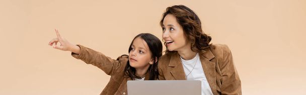 modna dziewczyna wskazując palcem w pobliżu laptopa i zdumiony mama odizolowany na beżowy, baner - Zdjęcie, obraz