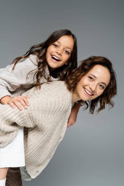 happy brunette woman in knitwear piggybacking cheerful daughter isolated on grey - Photo, image