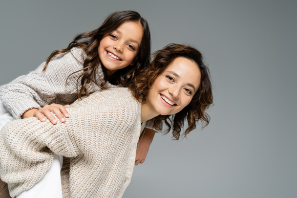 young woman in warm clothes piggybacking daughter while looking at camera isolated on grey - Photo, Image
