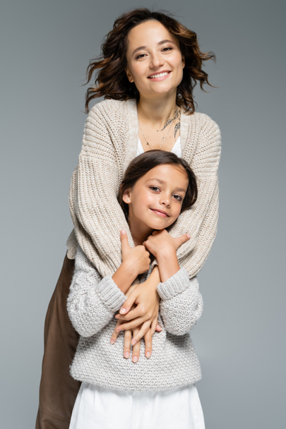 ενθουσιασμένοι μελαχρινή γυναίκα σε μοντέρνα πλεκτά αγκαλιάζει χαμογελαστή κόρη απομονωμένη σε γκρι - Φωτογραφία, εικόνα