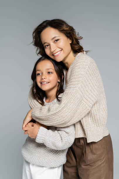 stylish brunette woman looking at camera while hugging daughter isolated on grey - Photo, Image
