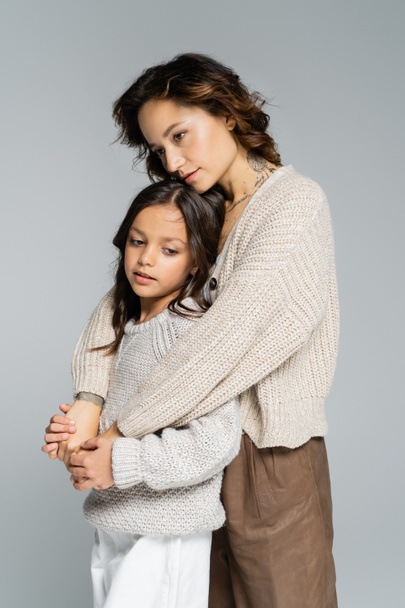 dreamy woman in trendy autumn clothes hugging daughter isolated on grey - Фото, изображение