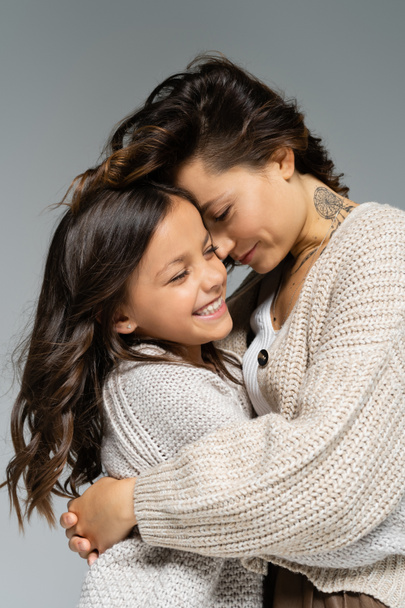 tattooed woman in warm clothes embracing excited daughter isolated on grey - Photo, Image