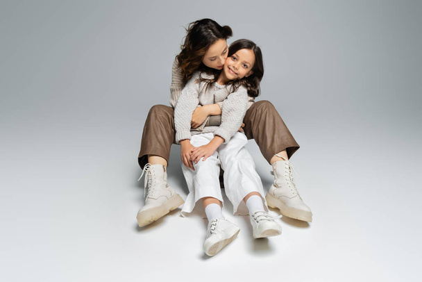 full length of stylish woman embracing daughter while sitting on grey background - Φωτογραφία, εικόνα