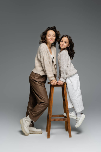 full length of mother and daughter in trendy autumn outfit posing near wooden stool on grey  - Фото, зображення