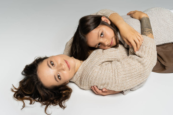 brunette mom and daughter in cozy knitwear smiling at camera and hugging on grey background - Foto, immagini