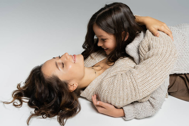 smiling woman in warm cardigan embracing daughter while lying on grey background - Фото, зображення