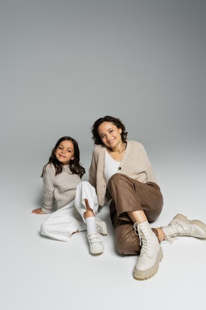 full length of mother and daughter in knitwear and pants sitting on grey background - Fotoğraf, Görsel