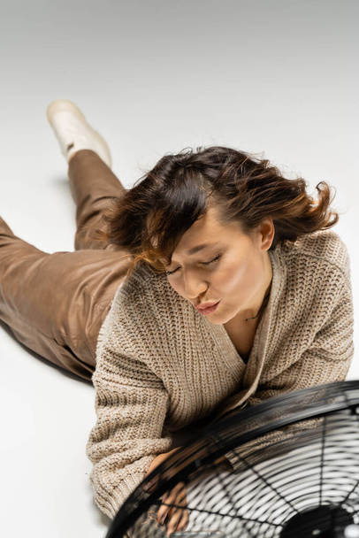 woman in warm cardigan pouting lips while lying near blowing electric fan on grey background - Foto, Imagem