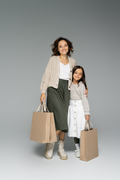 happy woman in stylish autumn clothes holding shopping bags and hugging daughter on grey background - Фото, зображення