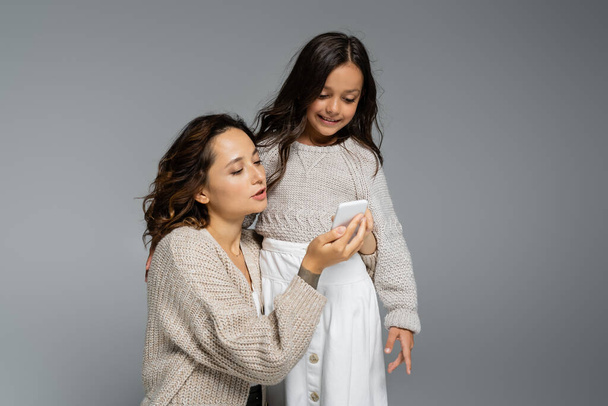 brunette woman showing mobile phone to smiling daughter isolated on grey - Foto, afbeelding