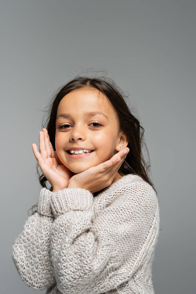 excited child in warm sweater holding hands near face and looking at camera isolated on grey - Foto, Imagem