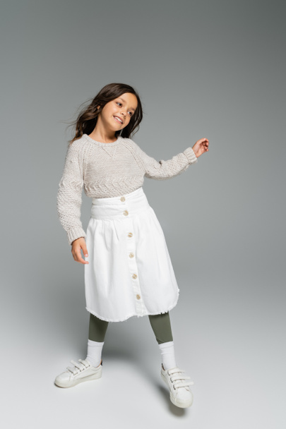 full length of happy girl in warm sweater and white skirt posing on grey background - Photo, Image