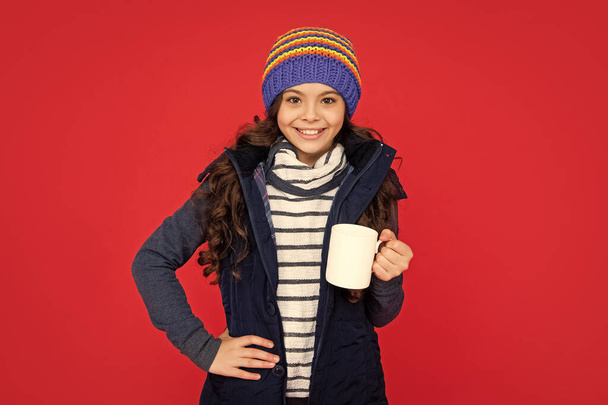 positive kid in puffer waistcoat and hat. teen girl drink warm tea on red background. portrait of child with coffee cup. drinking cocoa and milk. - Zdjęcie, obraz