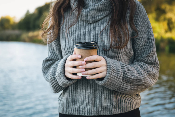 Close-up, uma xícara de café nas mãos de uma mulher na natureza junto ao rio, uma bebida takeaway na estação fria. - Foto, Imagem