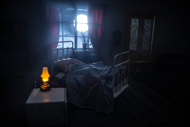 Horror-Halloween. Ein realistisches Puppenhaus-Schlafzimmer mit Möbeln und Fenster in der Nacht. Verängstigter Mann im Bett bei Vollmond draußen. Kreative Dekoration. Selektiver Fokus - Foto, Bild