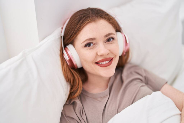 Young redhead woman listening to music lying on bed at bedroom - Фото, изображение