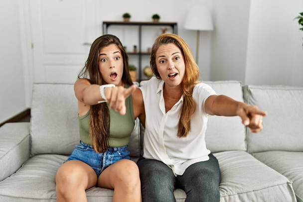 Mother and daughter together sitting on the sofa at home pointing with finger surprised ahead, open mouth amazed expression, something on the front  - Φωτογραφία, εικόνα