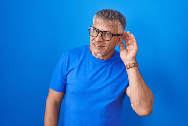 Homem hispânico com cabelos grisalhos em pé sobre fundo azul sorrindo com a mão sobre o ouvido ouvindo um rumor ou fofocas. conceito de surdez.  - Foto, Imagem