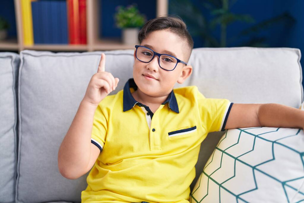 Joven niño hispano sentado en el sofá en casa sonriendo con una idea o pregunta apuntando con el dedo con la cara feliz, número uno  - Foto, Imagen