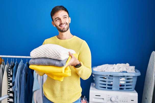 Young hispanic man smiling confident holding folded clothes at laundry room - Photo, Image