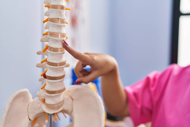 Jovem bela mulher hispânica fisioterapeuta apontando para modelo anatômico da coluna vertebral na clínica de reabilitação - Foto, Imagem