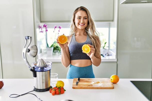Jovem mulher sorrindo confiante segurando laranja na cozinha - Foto, Imagem
