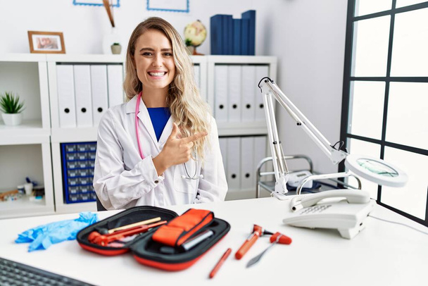 Молодая красивая женщина врач с рефлекторным молотком и медицинскими инструментами веселый с улыбкой лица указывая рукой и пальцем в сторону со счастливым и естественным выражением лица  - Фото, изображение
